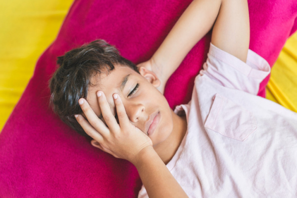 Brown-haired boy lying on colorful spread, looks pained, one hand covering an eye, other eye shut & other hand tucked behind head; concept is migraine headache
