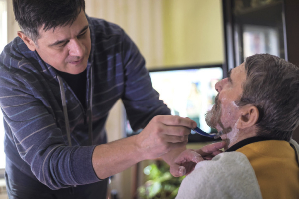 A home health aide wearing a blue striped shirt helping an older man, seated and wearing a yellow and black top, with his daily shave